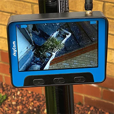 SkyCam monitor showing a live wireless view of a high-reach gutter being cleared of moss.